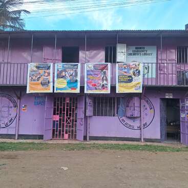 Un edificio morado de dos pisos alberga la Biblioteca Infantil Comunitaria Ubuntu, que cuenta con coloridos carteles y programas educativos, con niños cerca y una exuberante vegetación que rodea la zona.