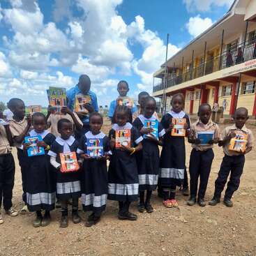 A group of school children stand outside holding new books and stationery, smiling. They wear uniforms and pose in front of a school building under a bright sky.