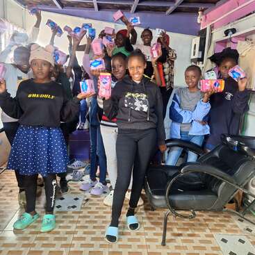 A group of smiling girls proudly hold up sanitary pads, standing together in a colorful room, celebrating access to menstrual products and supporting each other.