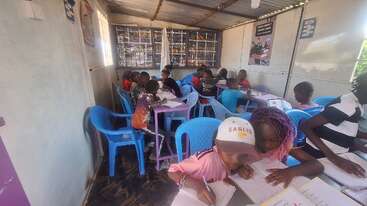 Children are seated in a classroom with blue plastic chairs, concentrating on their schoolwork. Bright sunlight illuminates the space, and educational posters decorate the walls.