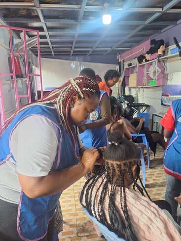 In a brightly lit salon, women wearing blue aprons are braiding clients' hair. The atmosphere is busy and collaborative, with hair products and wigs on shelves.
