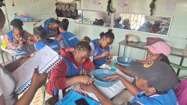 A group of women in blue vests practice nail care in a classroom setting, focused on hands-on learning with water bowls, notebooks, and various manicure tools.