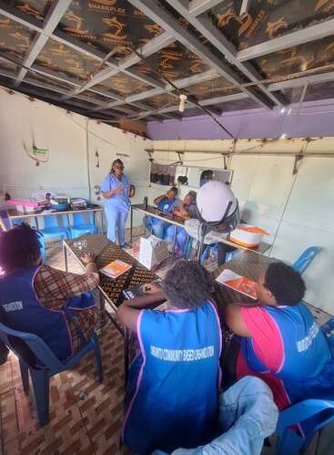 A group of people in blue "Ubuntu Community Based Organization" vests attend a training or meeting in a room, listening to a standing facilitator.