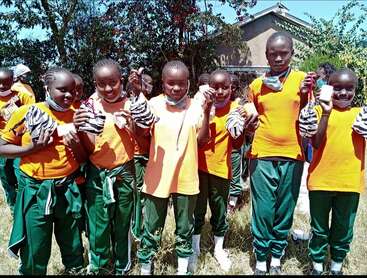 A group of children in orange shirts and green pants stand outdoors, smiling and holding striped cloth items and small white objects, likely gifts or supplies.