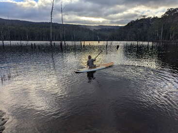 Eine Person gleitet auf einem Paddleboard über einen ruhigen, spiegelglatten See, der von Bäumen umgeben ist, unter einem bewölkten Himmel, durch den die Sonne scheint – eine friedliche Szene.