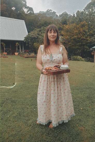 Eine Frau in einem geblümten Kleid steht barfuß auf dem Rasen und hält ein Holztablett mit Essen in der Hand. Im ruhigen Hintergrund sind Bäume und ein Haus zu sehen.