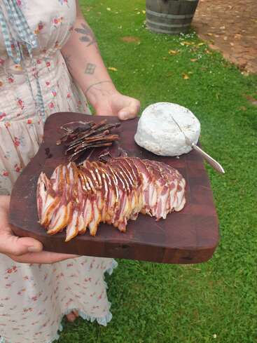 Eine Person in einem geblümten Kleid hält in einer grasbewachsenen Außenkulisse ein Holzbrett mit Aufschnitt, Biltong und einem runden Käse in der Hand.