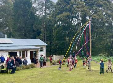 Vor einem Gemeindesaal tanzen Kinder mit bunten Bändern um einen Maibaum. Zuschauer verfolgen das Fest. Hohe Bäume und viel Grün umgeben das ländliche Beisammensein.