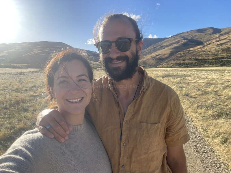 La imagen muestra a un hombre y una mujer juntos en un campo, con colinas onduladas y un cielo azul de fondo, en un día soleado.