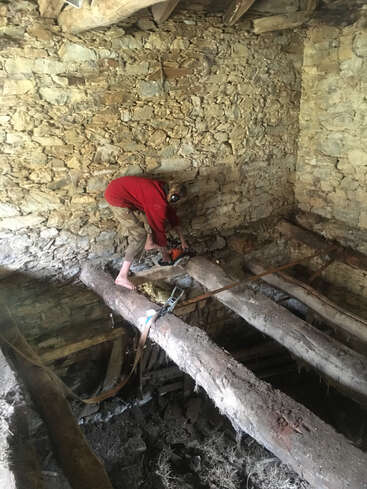 Ein Mann in rotem Hemd und khakifarbener Hose sägt mit einer Kettensäge einen Baumstamm in einem rustikalen Steinhaus mit freiliegenden Holzbalken.