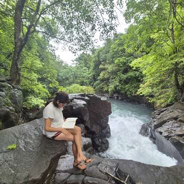 Eine Frau sitzt auf einem Felsen am Fluss, vertieft in ein Buch, umgeben von üppigem Grün und einer ruhigen natürlichen Umgebung.