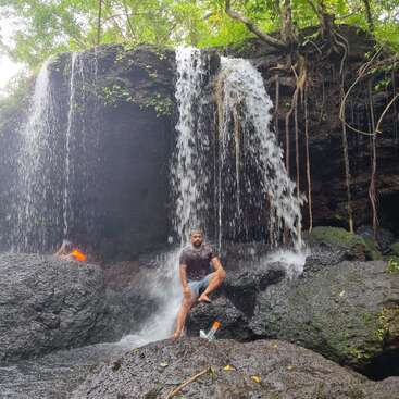 Ein Mann posiert auf dunklen Felsen unter einem fließenden Wasserfall, umgeben von üppigem Grün. Eine Wasserflasche liegt in der Nähe, und eine weitere Person ist teilweise im Hintergrund zu sehen.