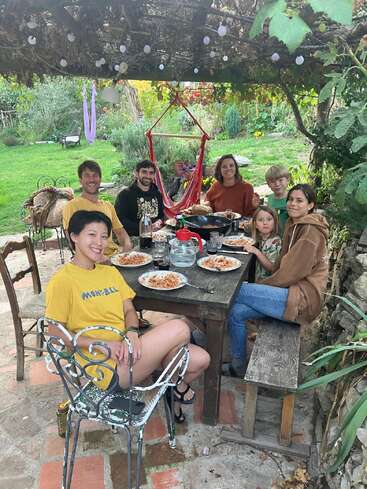Seis personas disfrutan de una comida de espaguetis al aire libre, sentadas en una mesa rústica de madera bajo unas parras, sonrientes, rodeadas de exuberante vegetación y en un acogedor entorno ajardinado.