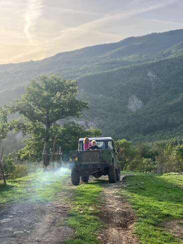 Dos niños viajan en la parte trasera de una vieja camioneta verde por un camino de tierra, rodeados de exuberantes colinas verdes y un paisaje tranquilo y pintoresco.
