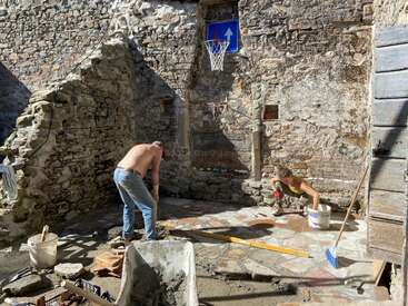 Dos personas trabajan en la restauración de un patio de piedra bajo el sol. Hay herramientas y materiales de construcción esparcidos por los alrededores. Una canasta de baloncesto y un cartel adornan el viejo muro.