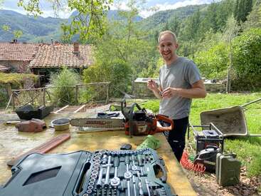 Un hombre sonríe al aire libre junto a una mesa con herramientas, una motosierra y equipamiento, sobre un fondo de árboles verdes y casas rústicas con tejados rojos.