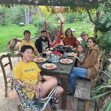 Six people enjoy a meal of spaghetti outdoors, seated at a rustic wooden table under vines, smiling, surrounded by lush greenery and a cozy garden setting.