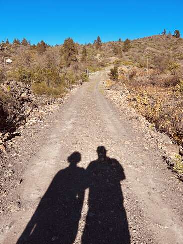 A sunny, clear day on a rocky dirt path through hilly terrain. Two people’s shadows, close together, are cast onto the road amid surrounding bushes.