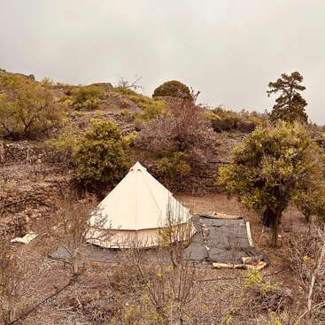 A beige canvas tent sits amidst dry shrubs and scattered trees on rugged, terraced terrain under a cloudy sky, creating a serene and rustic camping scene.