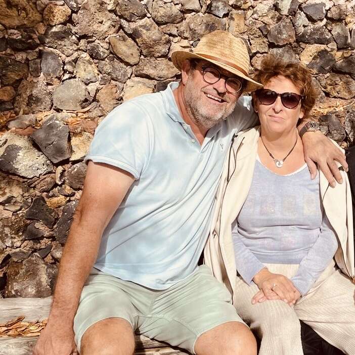 Un hombre sonriente con sombrero y gafas está sentado con el brazo alrededor de una mujer que lleva gafas de sol. Están sentados juntos en un banco junto a un muro de piedra.