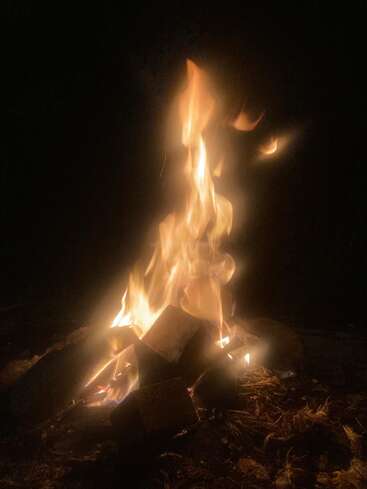 Uma fogueira brilhante arde na escuridão, com as chamas dançando sobre os troncos de madeira. O brilho quente ilumina os arredores, criando uma atmosfera aconchegante e convidativa na noite.