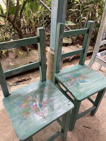 Duas cadeiras de madeira verdes com padrões florais pintados nos assentos estão colocadas do lado de fora, perto de uma cerca, cercadas por plantas e elementos naturais ao ar livre.