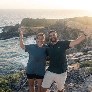 Das Bild zeigt einen Mann und eine Frau, die auf einer felsigen Klippe mit Blick auf das Meer stehen und ihre Arme in einer freudigen Pose heben.