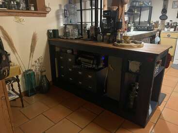 A cozy kitchen scene with a wooden island, coffee maker, jars, water dispenser, and decorative vases. Warm lighting and organized shelves create a homely atmosphere.