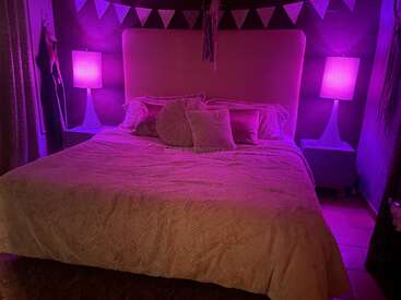 A cozy bedroom with a large bed, pink and purple lighting, two lamps on nightstands, decorative pillows, and a triangular flag bunting hanging above the headboard.