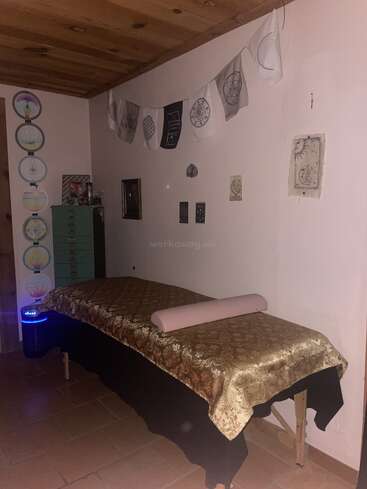 A tranquil massage room with a wooden ceiling, decorated walls, golden embroidered tablecloth, pink bolster, mandala art, prayer flags, and a calming ambiance.