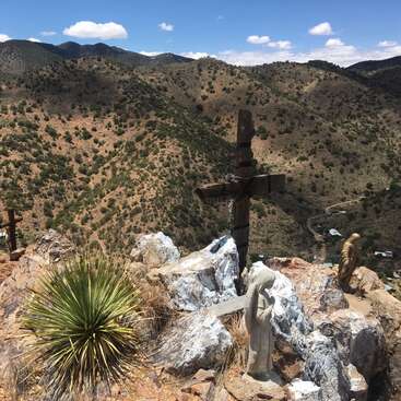 A rugged desert mountain landscape features wooden crosses, religious statues, white rocks, and a spiky yucca plant under a bright blue sky with scattered clouds.