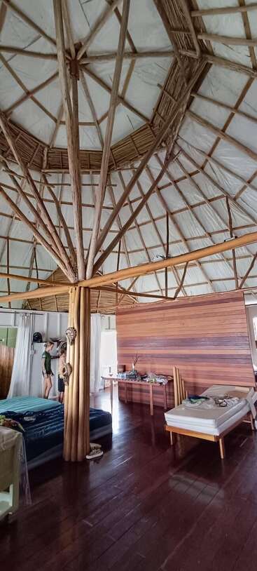 Une pièce spacieuse et rustique avec un haut plafond rond soutenu par des poutres apparentes. On y voit deux lits, des meubles en bois et des personnes, ce qui lui confère une atmosphère chaleureuse et écologique.