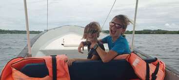 Deux enfants aux cheveux ébouriffés par le vent rient joyeusement sur un bateau, portant des lunettes de soleil. Des gilets de sauvetage orange vif les entourent. L'arrière-plan montre de l'eau et un ciel nuageux.