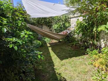 Une personne se détend dans un hamac, entourée d'une végétation luxuriante, dans un jardin intime. Un auvent blanc offre de l'ombre sous un ciel d'un bleu éclatant.