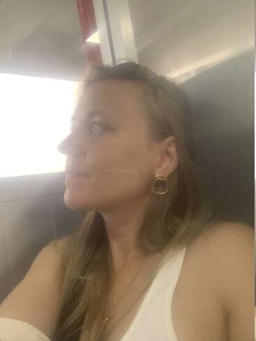 Une femme aux longs cheveux blonds est assise à l'intérieur, le regard pensif tourné vers sa gauche. Elle porte des boucles d'oreilles en or et un haut blanc, baignée dans une lumière douce.
