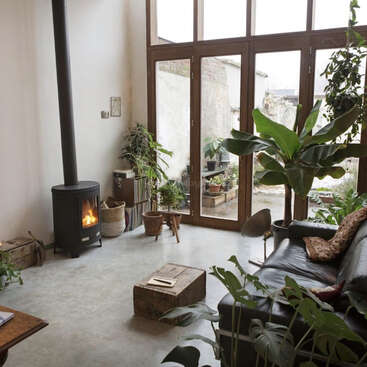 Un salon chaleureux équipé d'un poêle à bois, de nombreuses plantes en pot, de grandes baies vitrées, d'un canapé en cuir, d'une table basse rustique en bois et baigné de lumière naturelle.