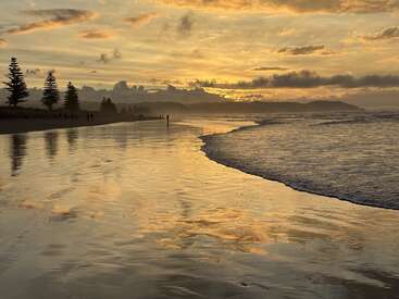 Der goldene Sonnenuntergang spiegelt sich wunderschön an einem ruhigen Strand. Silhouetten von Bäumen und Menschen in der Ferne unterstreichen die friedliche Atmosphäre. Sanfte Wellen gehen nahtlos in den leuchtenden Himmel über.