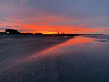 Ein ruhiger Strand bei Sonnenuntergang, ein leuchtend orangefarbener und rosafarbener Himmel, der sich auf dem nassen Sand spiegelt, entfernte Silhouetten von Bäumen und ruhige Wellen, die eine friedliche und magische Atmosphäre schaffen.