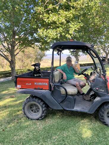 A man sits relaxed in a Kubota utility vehicle, wearing sunglasses and a hat. A happy dog is in the back. Trees and fields surround them.