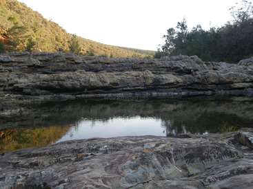 La imagen representa un sereno paisaje rocoso con una tranquila masa de agua, rodeada de exuberante vegetación y una ladera al fondo, bajo un cielo luminoso.