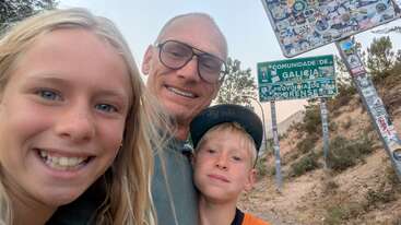 A smiling family selfie with a father and two children, set outdoors near a hillside. Behind them are Spanish road signs for Galicia and Ourense, covered in stickers.