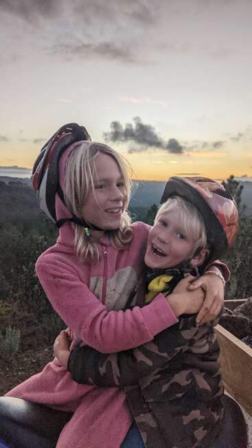 The image depicts two children, a girl and a boy, embracing in front of a scenic backdrop, with the girl wearing a pink hoodie and the boy sporting a red helmet.