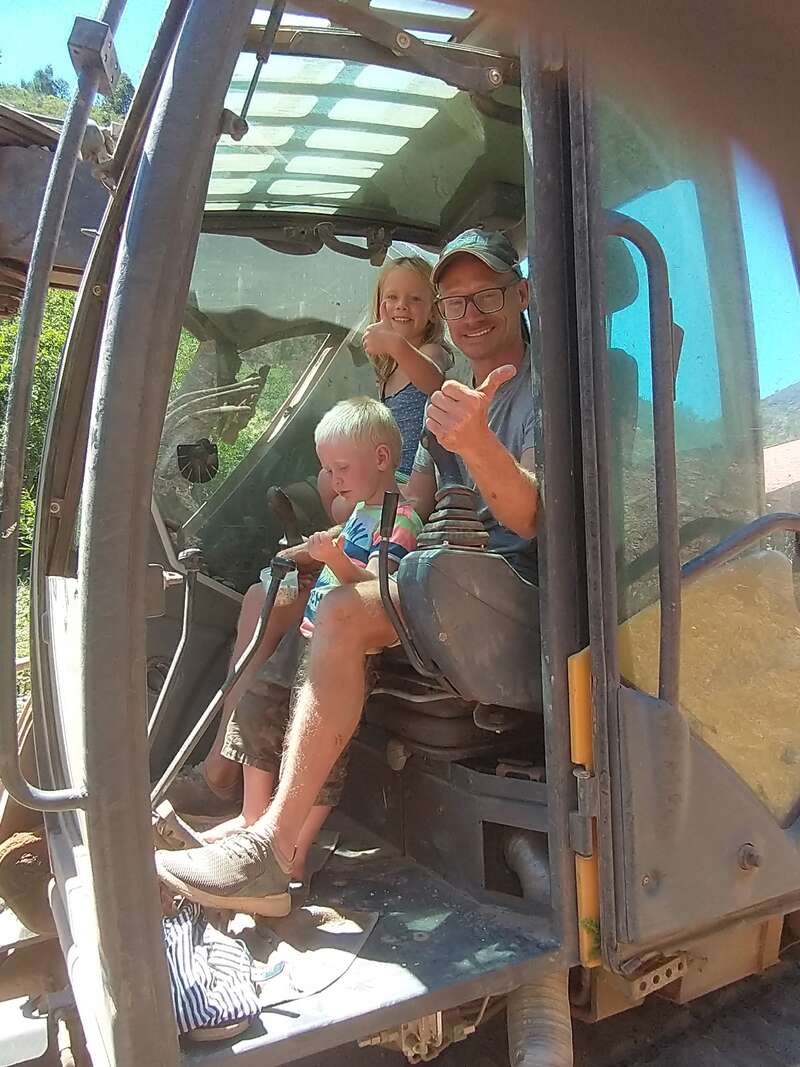 The image shows a man and two children sitting in a large vehicle, likely a tractor or excavator, with the man giving a thumbs-up.