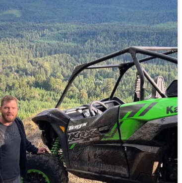 A man stands beside a green and black off-road vehicle on a scenic mountain overlook, surrounded by dense forest and rolling hills under a clear sky.