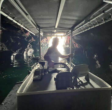 A person steers a boat inside a dark cave, heading towards the bright entrance. Rocky cave walls reflect on the water, creating a mysterious, adventurous atmosphere.
