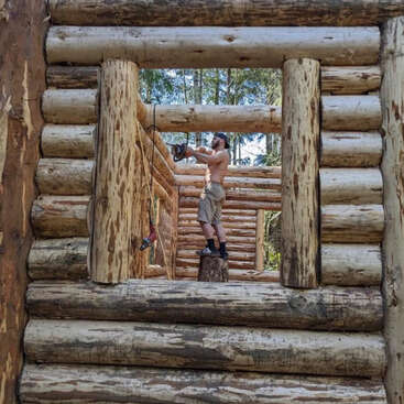 A shirtless man stands on a tree stump inside an unfinished log cabin, using a chainsaw. Sunlight filters through trees, highlighting the rustic wooden structure.