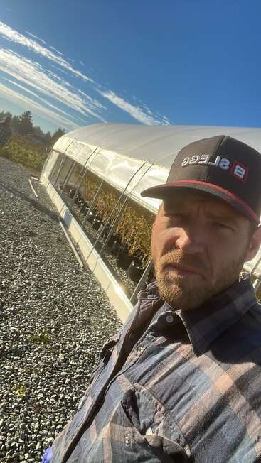 A man in a plaid shirt and black cap stands outdoors beside a greenhouse filled with plants on a gravel path under a clear blue sky.