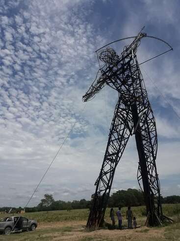 Una imponente escultura metálica de un arquero se alza en un campo, eclipsando a las personas cercanas y a un camión aparcado bajo un cielo azul parcialmente nublado. Majestuosa e impresionante.