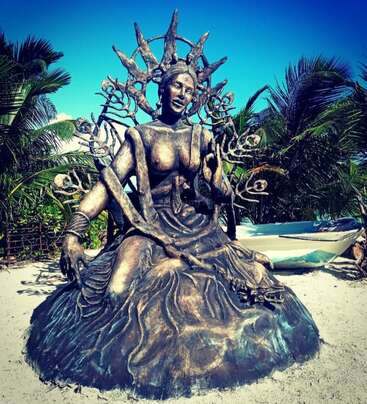 Una estatua de bronce de una mujer serena se sienta majestuosamente sobre la arena, adornada con una elaborada corona y formas orgánicas. Detrás de ella hay palmeras y una barca.