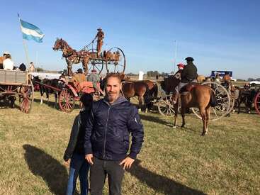 Un hombre frente a carruajes tradicionales tirados por caballos, personas vestidas de época, una bandera argentina y un campo bajo un cielo azul despejado.